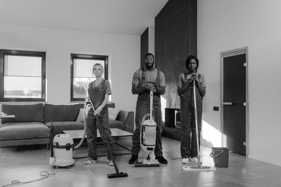 A group of three professional cleaners in a modern living room engaged in surface cleaning and deep cleaning tasks. The room features a light-colored wooden floor, a large sofa with cushions, and two 