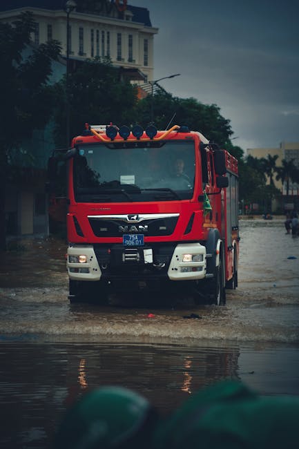 A bright red fire truck is positioned on a flooded urban street during a heavy rainstorm in Croydon. The vehicle is equipped with emergency lights on the roof and is facing forward across a waterlogge