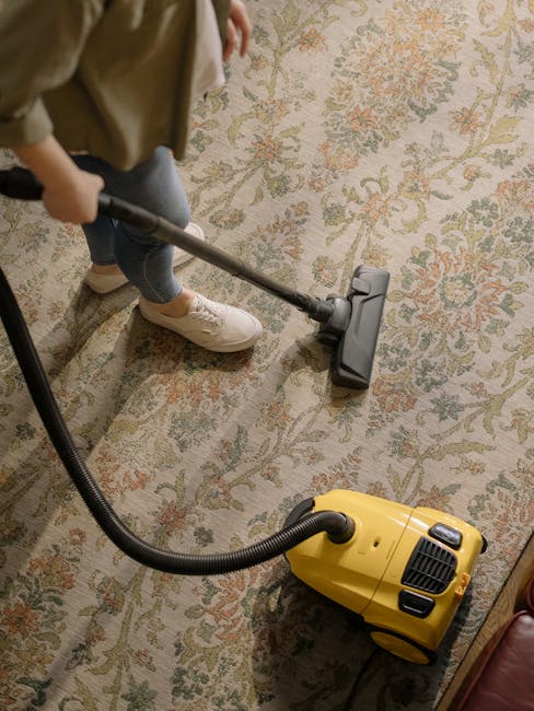 A yellow vacuum cleaner with a hose attachment is positioned on a decorative area rug in a room with wooden flooring. The vacuum is turned off and resting near a dark-colored TV stand that holds a whi