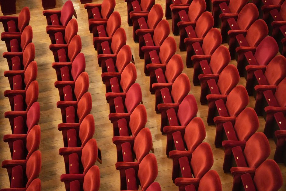 Interior view of a cinema or theatre auditorium with multiple rows of red upholstered seats featuring black armrests and headrests, arranged in a tiered layout. The seats are clean and well-maintained